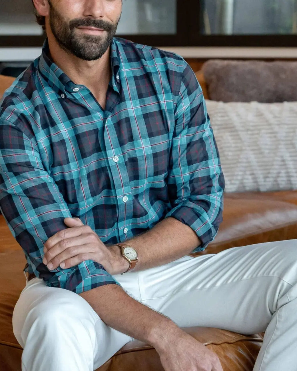 Man wearing Ledbury Blue Albini Olson Plaid shirt seated on leather sofa, paired with white chinos, sleeves rolled