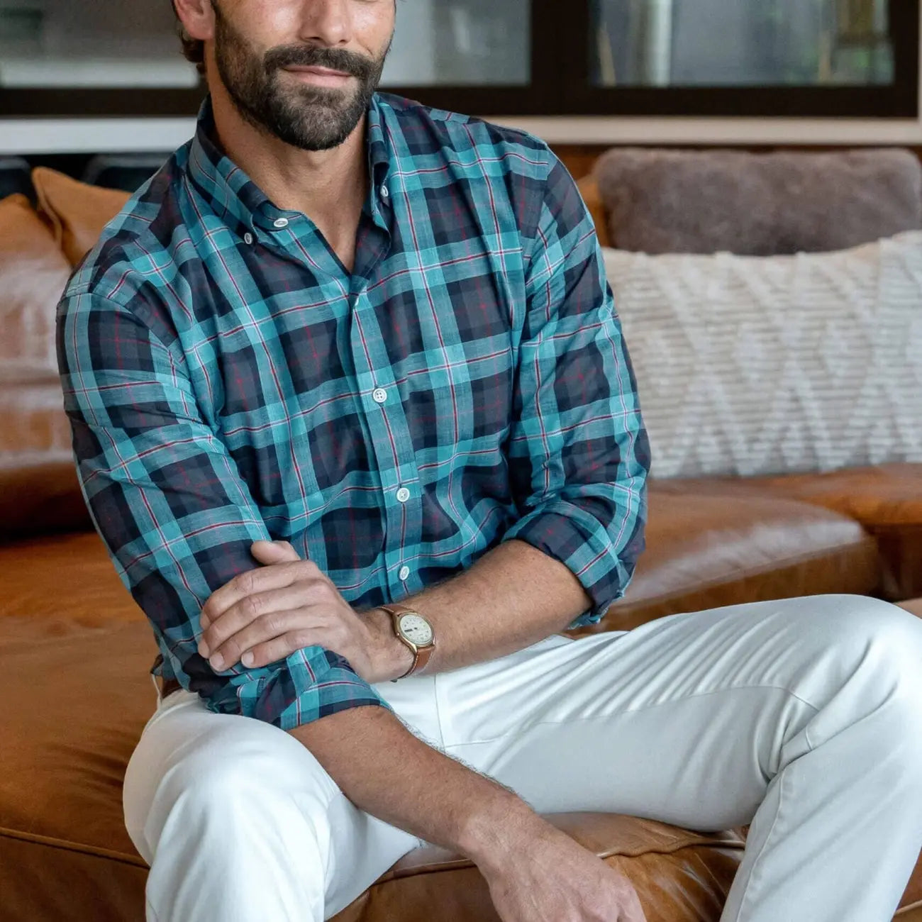 Man wearing Ledbury Blue Albini Olson Plaid shirt seated on leather sofa, paired with white chinos, sleeves rolled