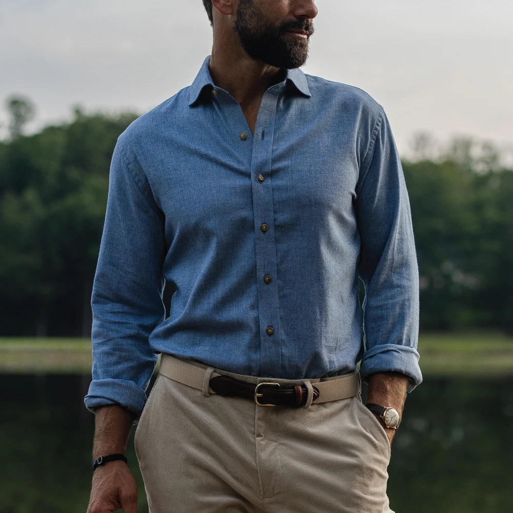 A man wearing a blue long-sleeve button-down shirt and beige trousers, standing outdoors near a body of water with trees in the background.