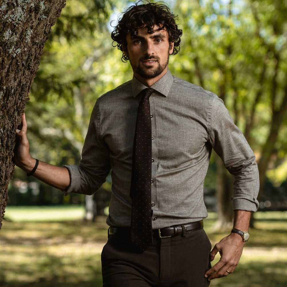 A man wearing a light brown long-sleeve button-down shirt, a dark patterned tie, and dark brown trousers, standing outdoors next to a tree in a park-like setting.