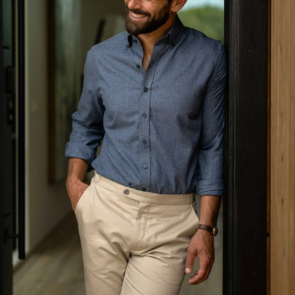 A man wearing a blue long-sleeve button-down shirt and beige trousers, standing in a doorway.