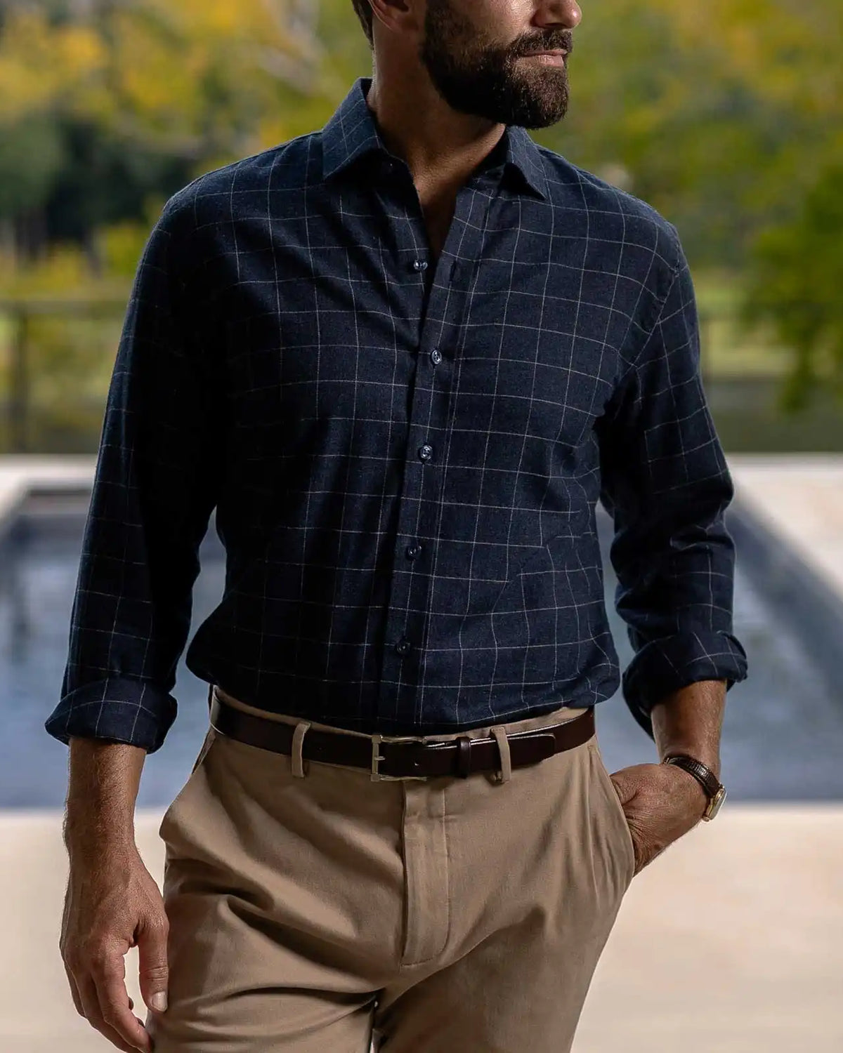 A man wearing a dark navy blue long-sleeve windowpane check button-down shirt tucked into khaki trousers, standing outdoors near a pool with a blurred green background.