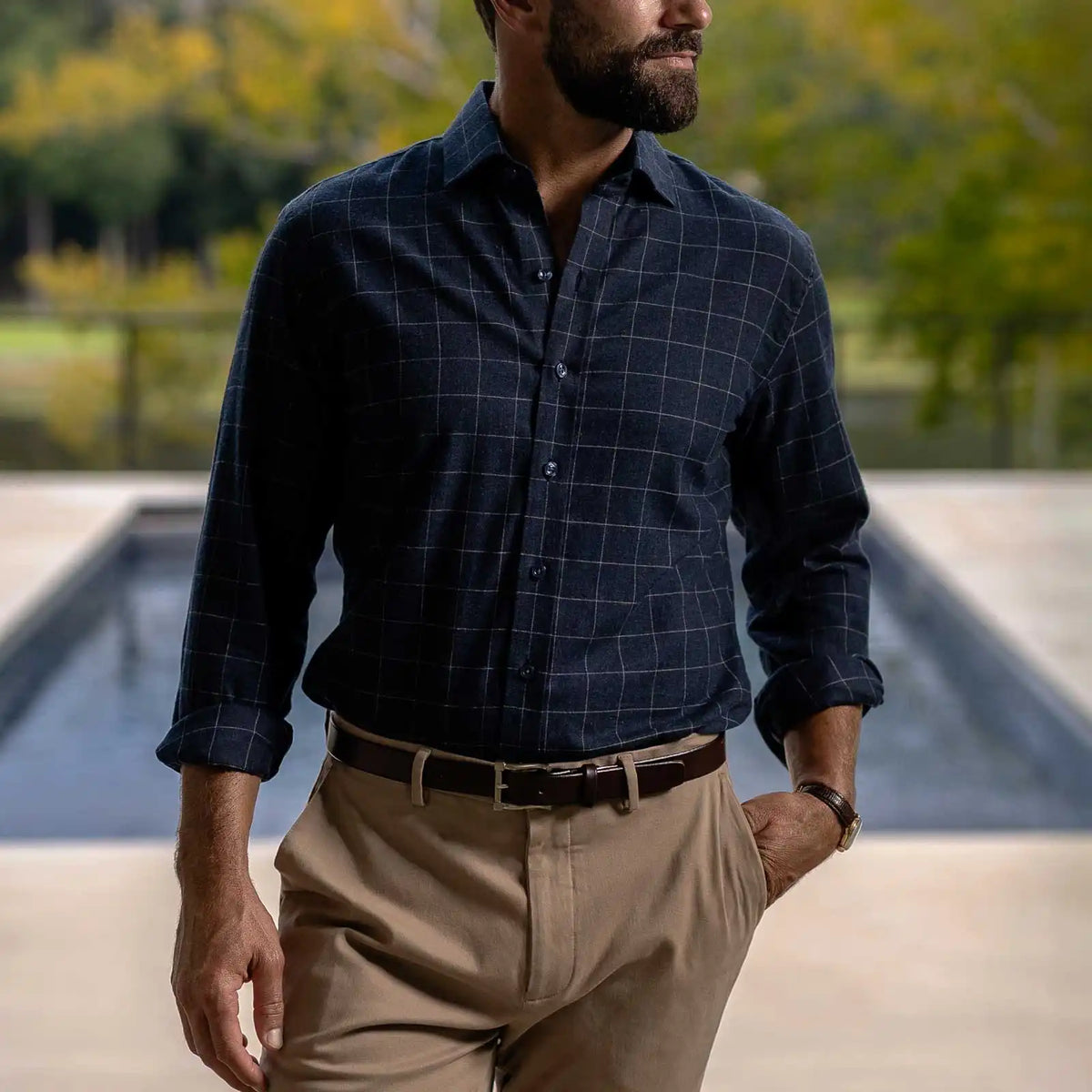 A man wearing a dark navy blue long-sleeve windowpane check button-down shirt tucked into khaki trousers, standing outdoors near a pool with a blurred green background.
