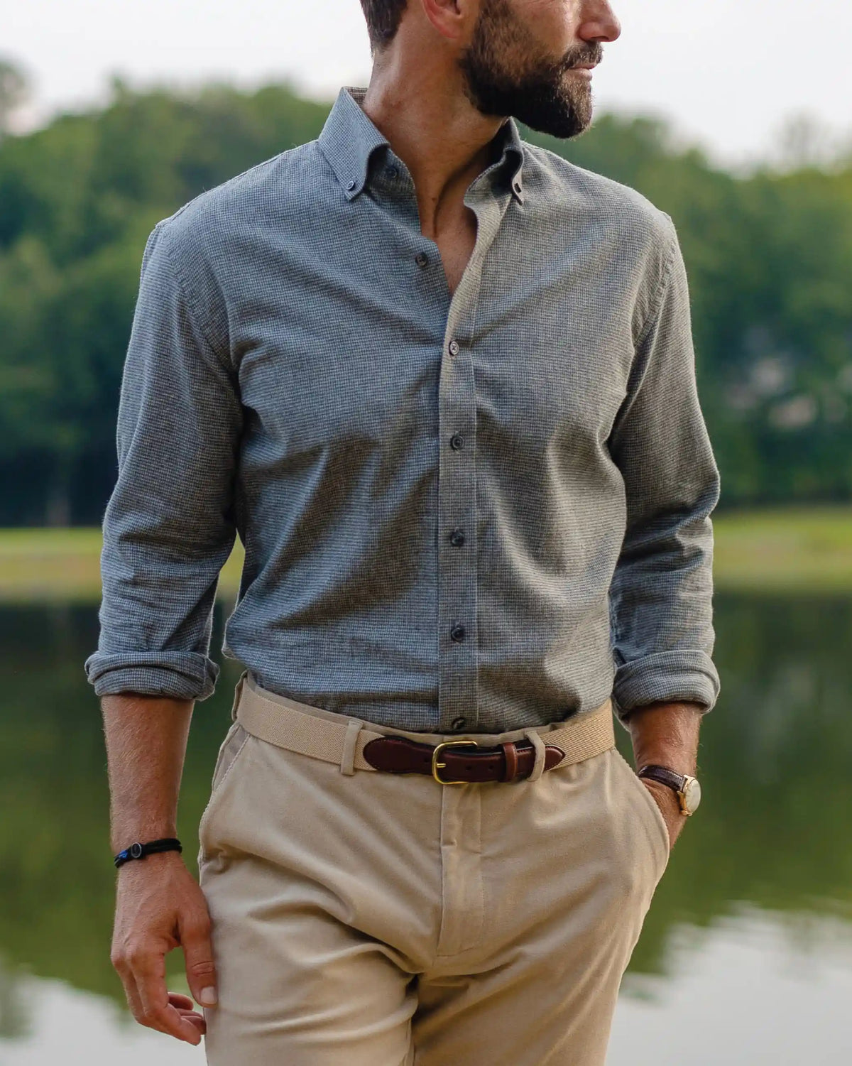 A man wearing a houndstooth grey long-sleeve button-down shirt and beige trousers, standing outdoors near a lake with trees in the background.