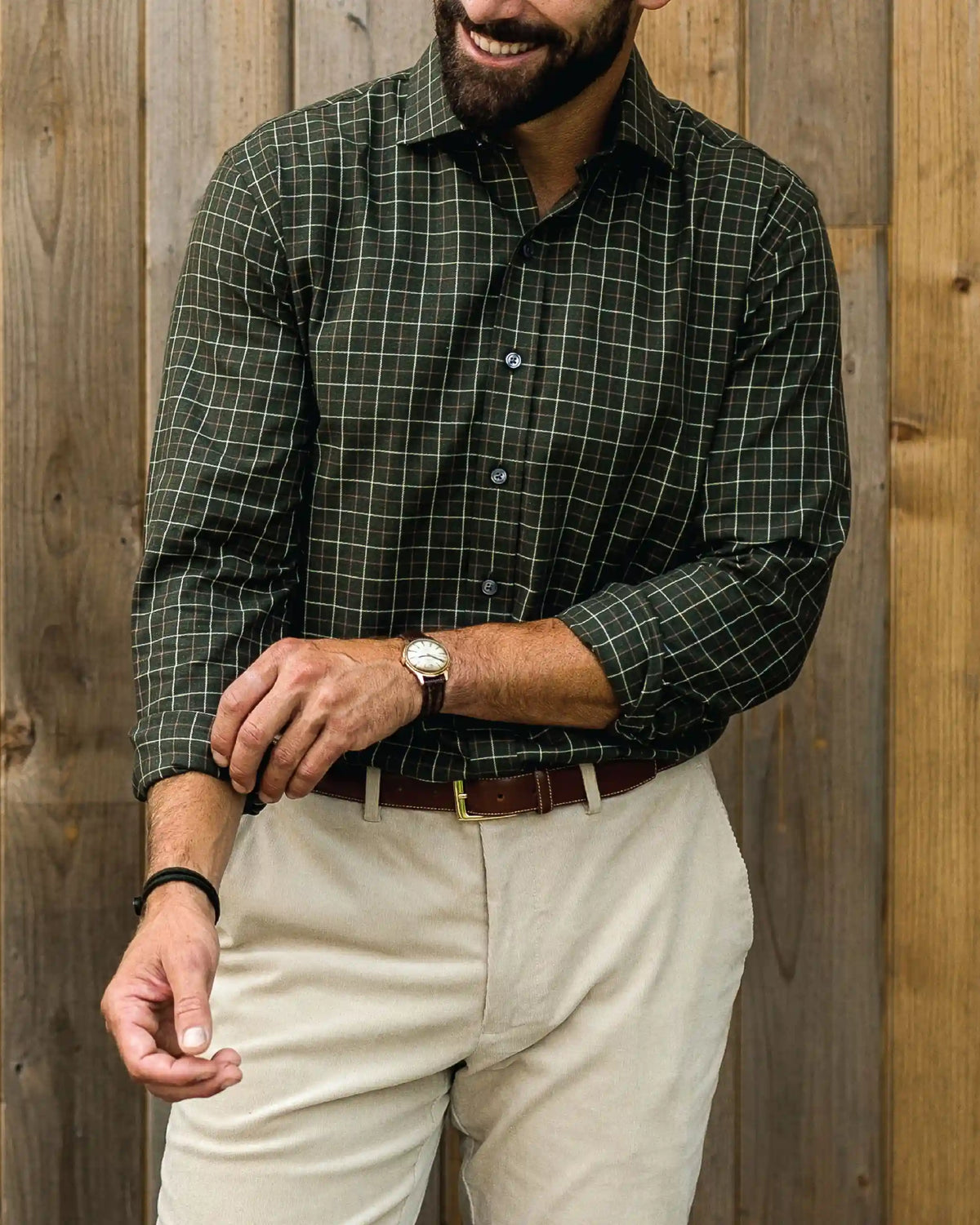A man wearing a dark green and black plaid long-sleeve button-down shirt with beige pants, standing in front of a vertical wooden plank wall.