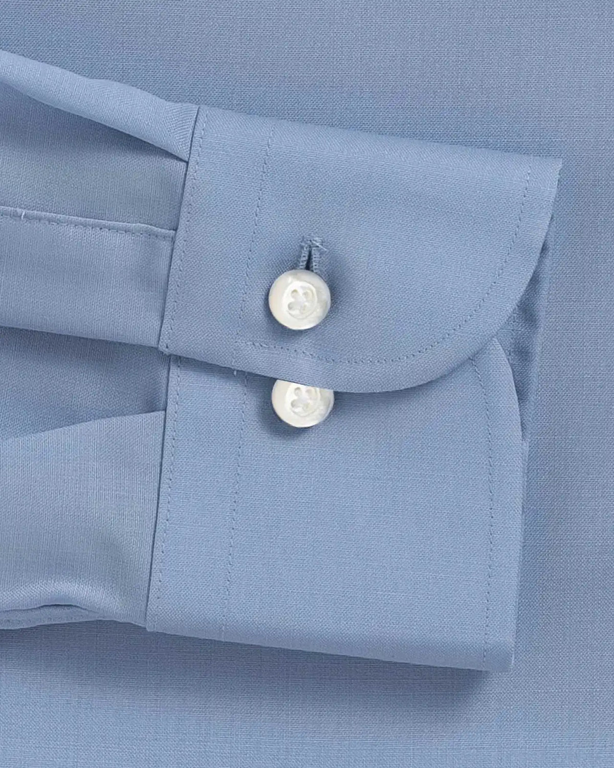 Close-up of a light blue dress shirt cuff with two white mother of pearl buttons on a matching blue fabric background.