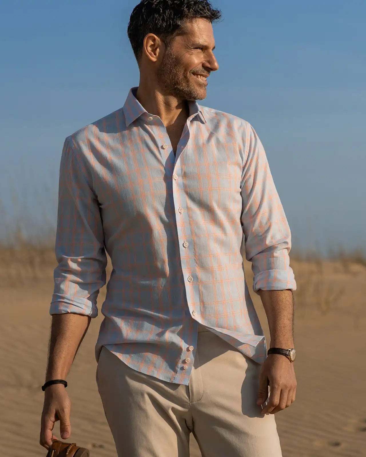 Man standing on a beach wearing a light blue and bright orange checkered shirt and beige pants.