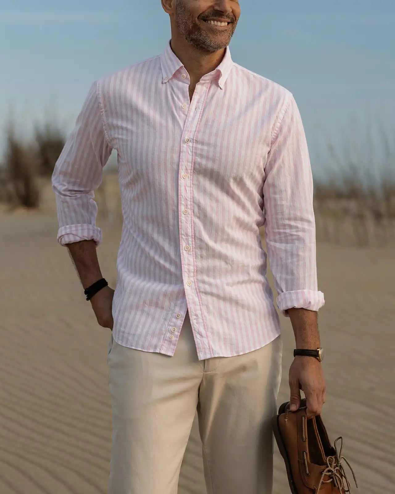 Man in a pink striped shirt and beige pants standing on sand with a clear sky.