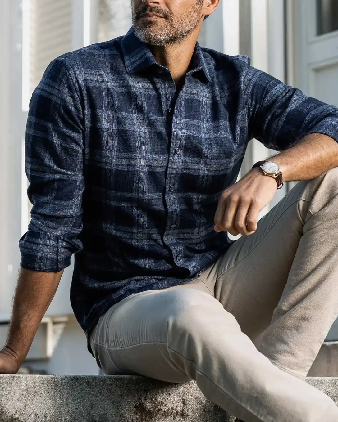A man wearing a navy blue and gray plaid button-down shirt with beige pants, sitting on concrete steps in front of a white building.