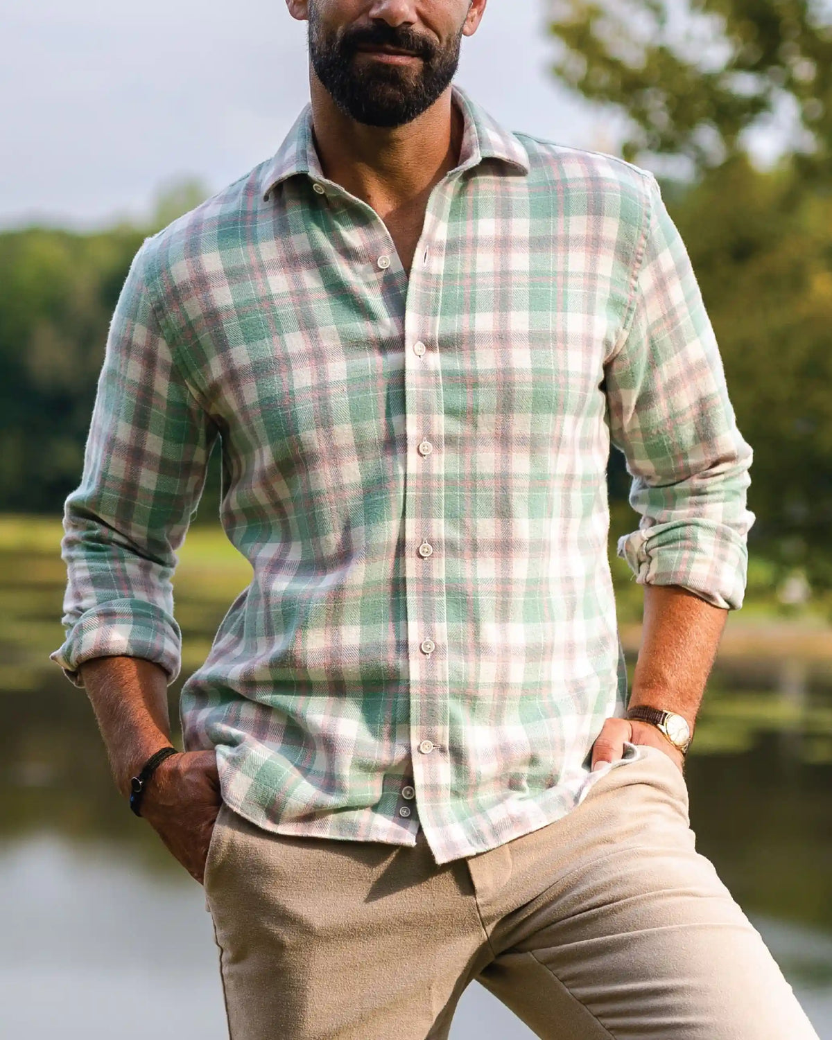 A man wearing a light green and beige plaid long-sleeve button-down shirt, paired with khaki pants, standing outdoors near a body of water with trees in the background.