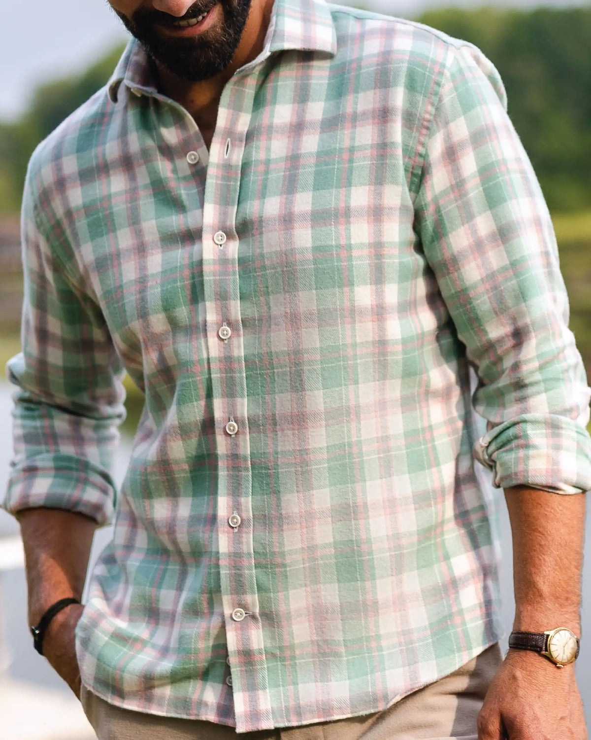 A man wearing a long-sleeve button-up plaid shirt in sage green, pink, and white, with the background showing a blurred outdoor scene of water and trees.