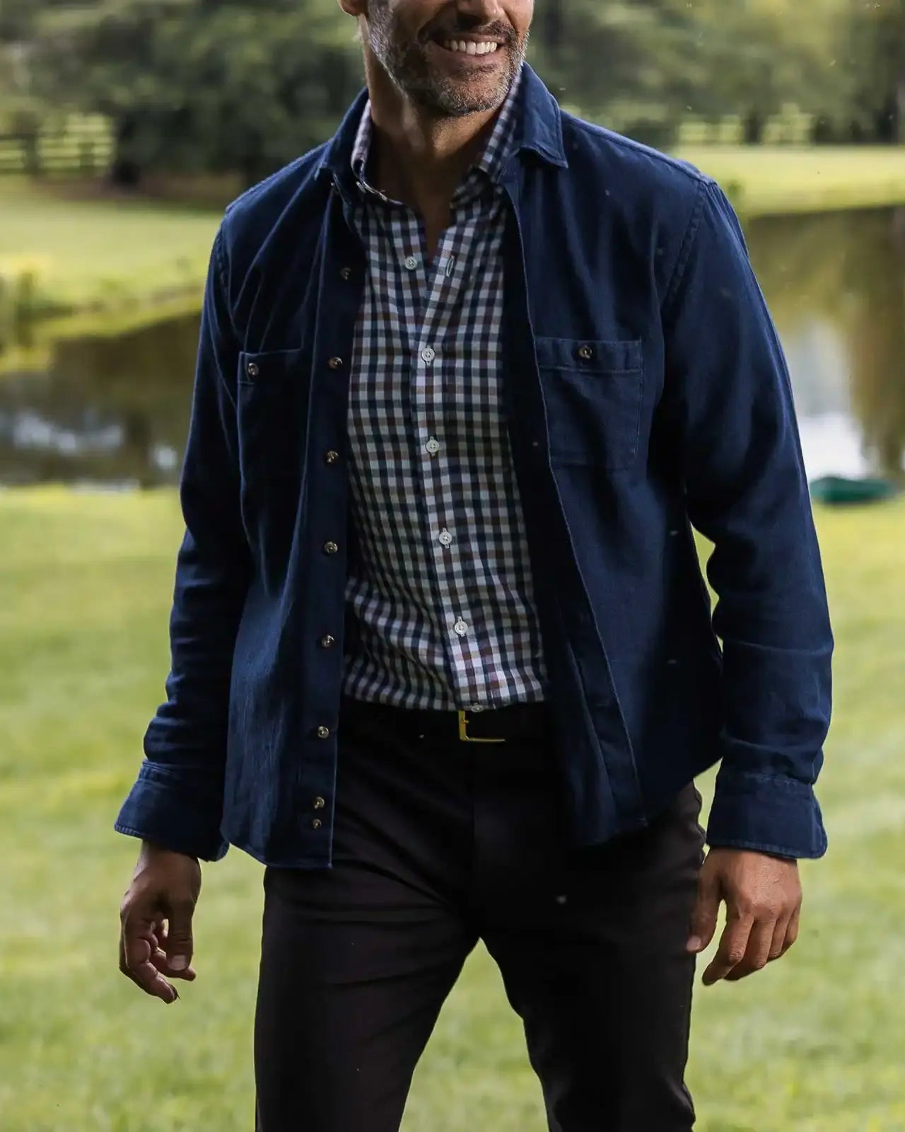 Man wearing a the blue Lisden utility shirt and checkered shirt standing in a park with a lake in the background