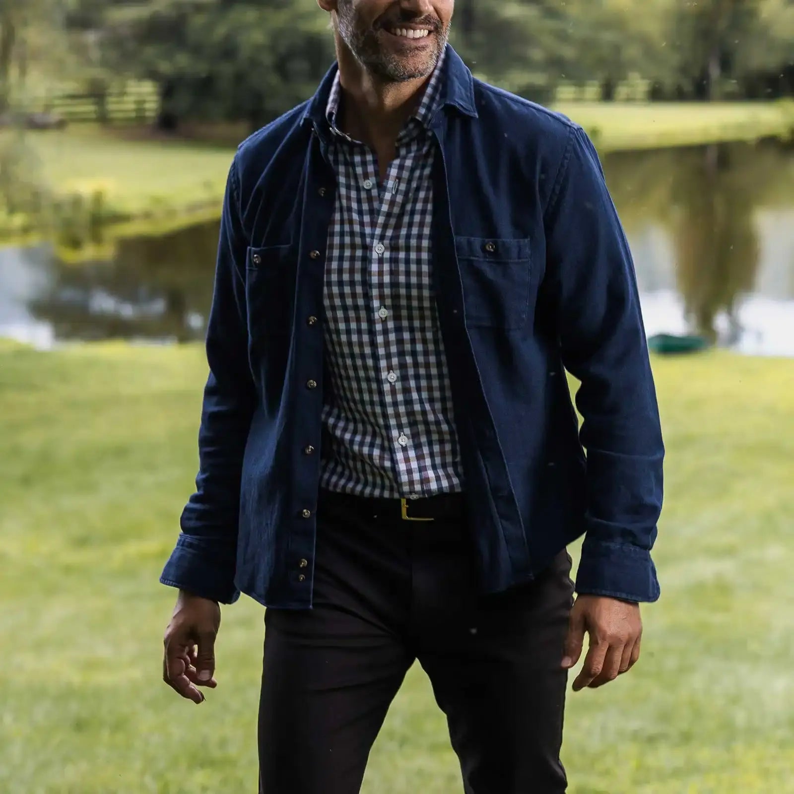 Man wearing a the blue Lisden utility shirt and checkered shirt standing in a park with a lake in the background