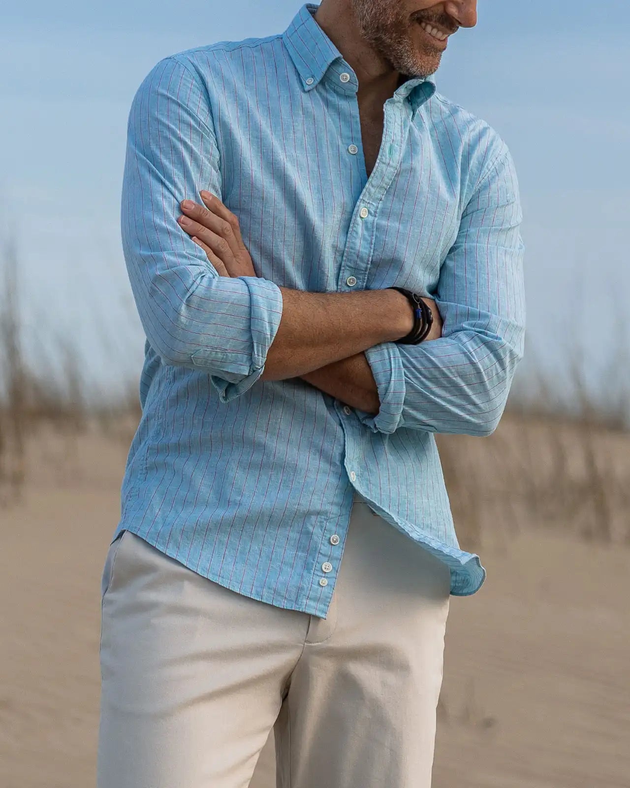 Man wearing a sea green and light red linen striped shirt and beige pants standing on a beach.