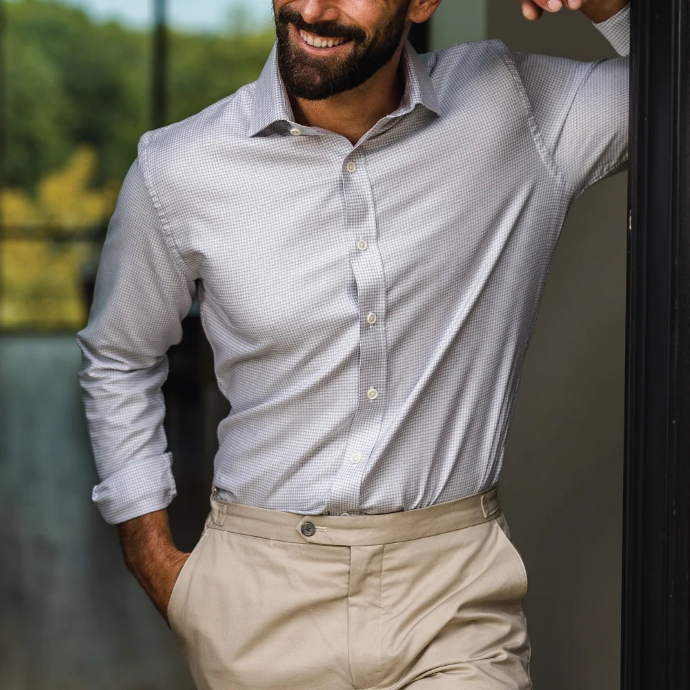 A man wearing a light gray and white micro-check patterned button-down dress shirt with beige trousers, standing in a doorway.