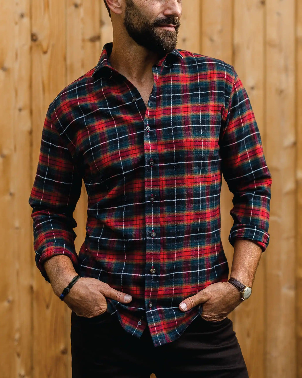 A man wearing a red, black, and white plaid long-sleeve button-down shirt, standing against a vertical wooden plank wall.