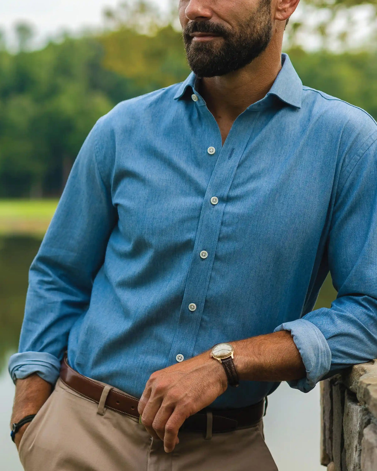 A man wearing a blue long-sleeve button-down shirt and khaki pants, leaning against a stone wall outdoors near water and trees.