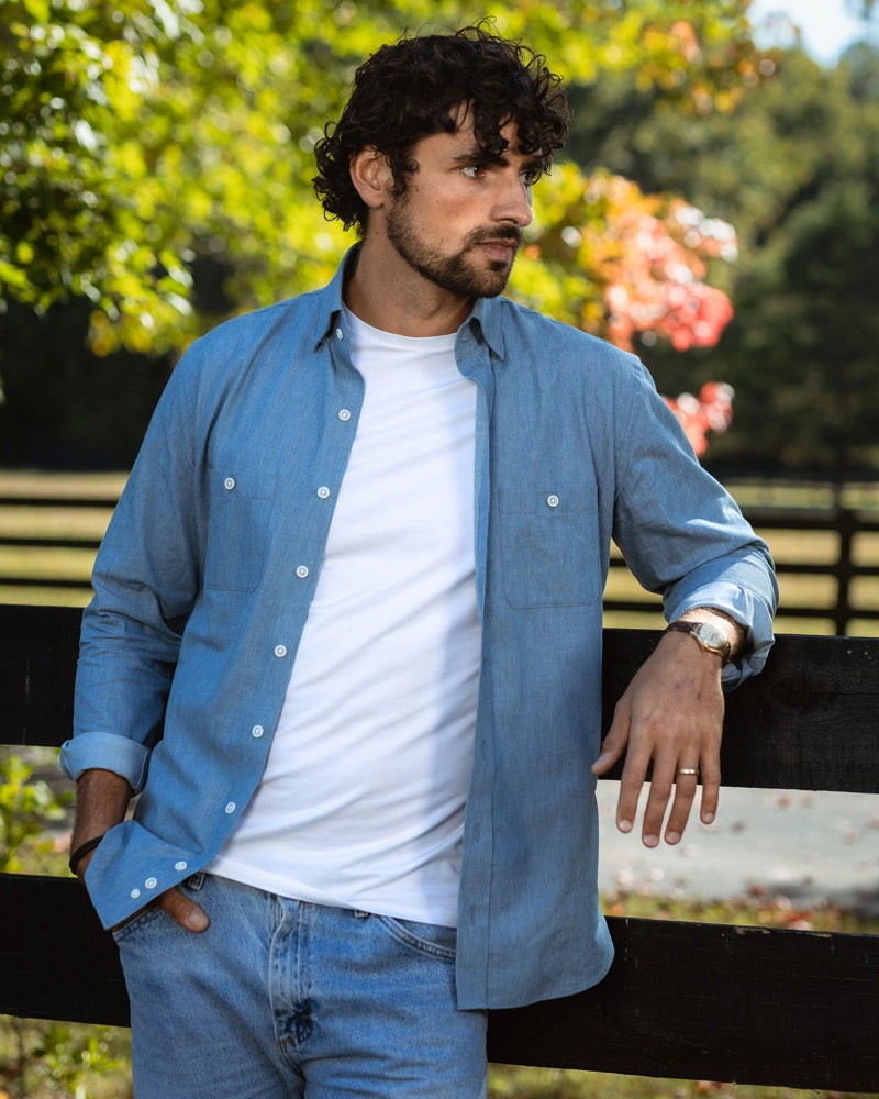 A man wearing a light blue button-down shirt over a white t-shirt and blue jeans, leaning on a black wooden fence in an outdoor park setting with green trees in the background.