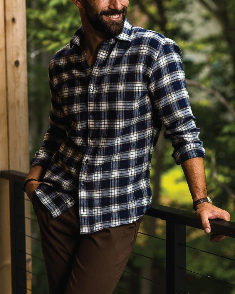 A man wearing a navy blue, white, and gray plaid button-down shirt and brown pants, leaning on a black metal railing on a wooden deck with a forest background.