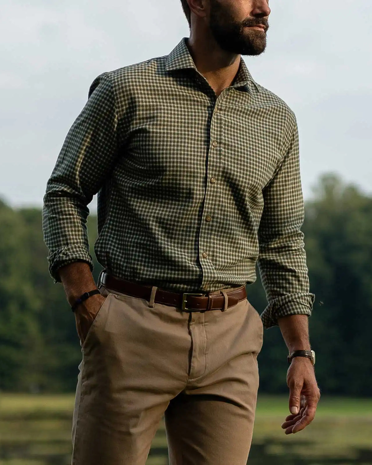A man wearing a green checkered long-sleeve button-down shirt, khaki pants, and a brown belt, standing outdoors in a grassy field with trees in the background.