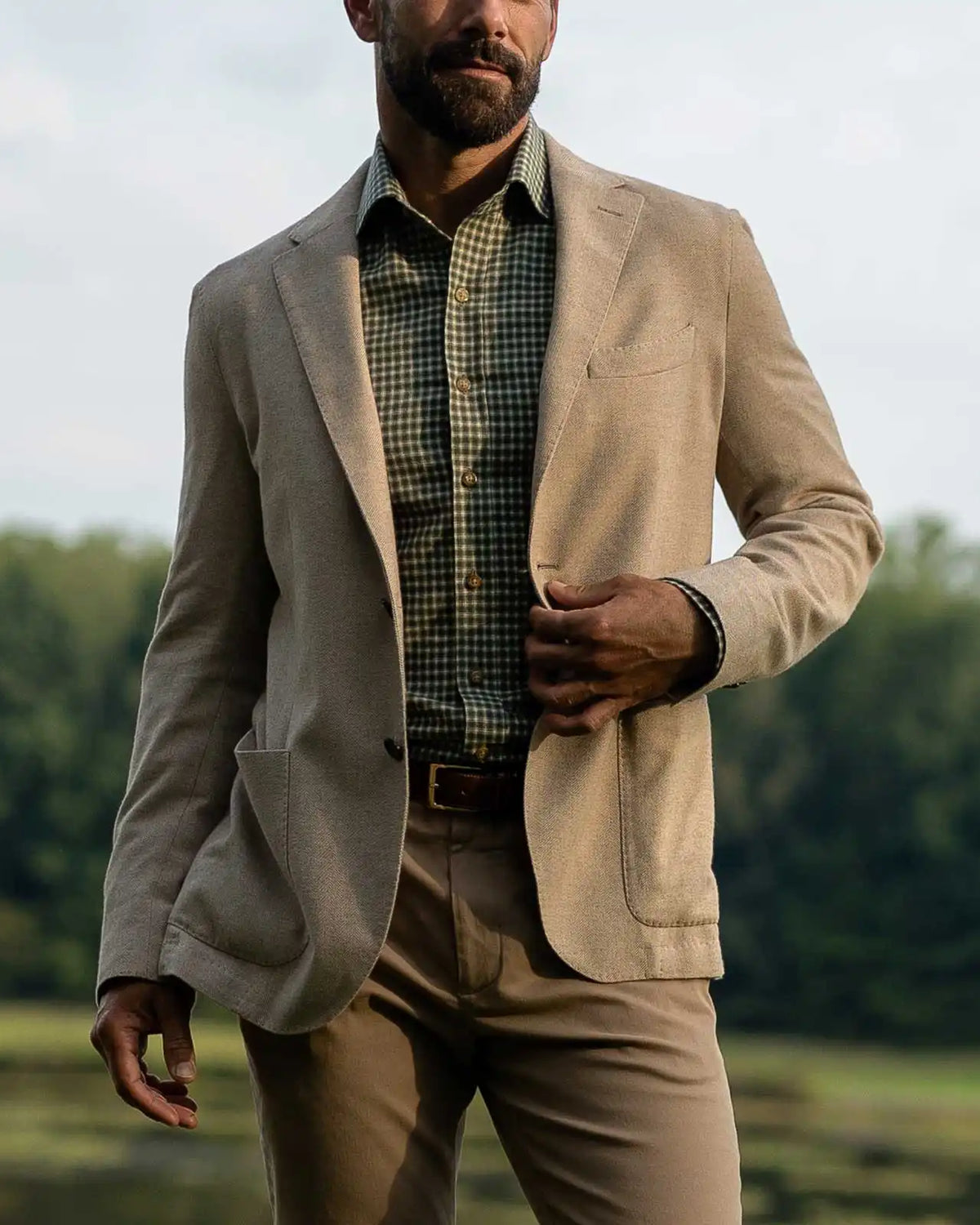 A man wearing a light beige blazer over a green checkered shirt, paired with matching beige trousers, standing outdoors in front of a blurred green landscape.