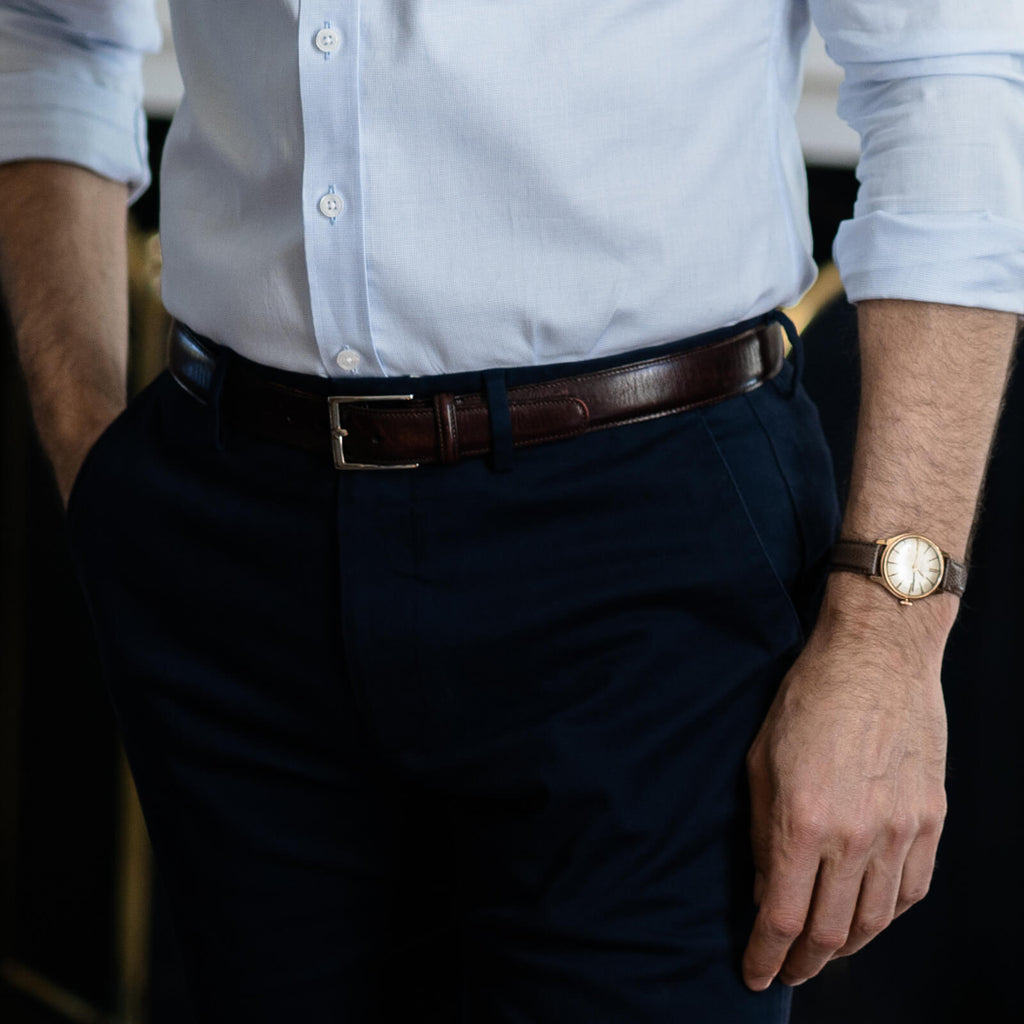 Closeup of a men's leather belt worn by a male model standing in front of a fireplace.