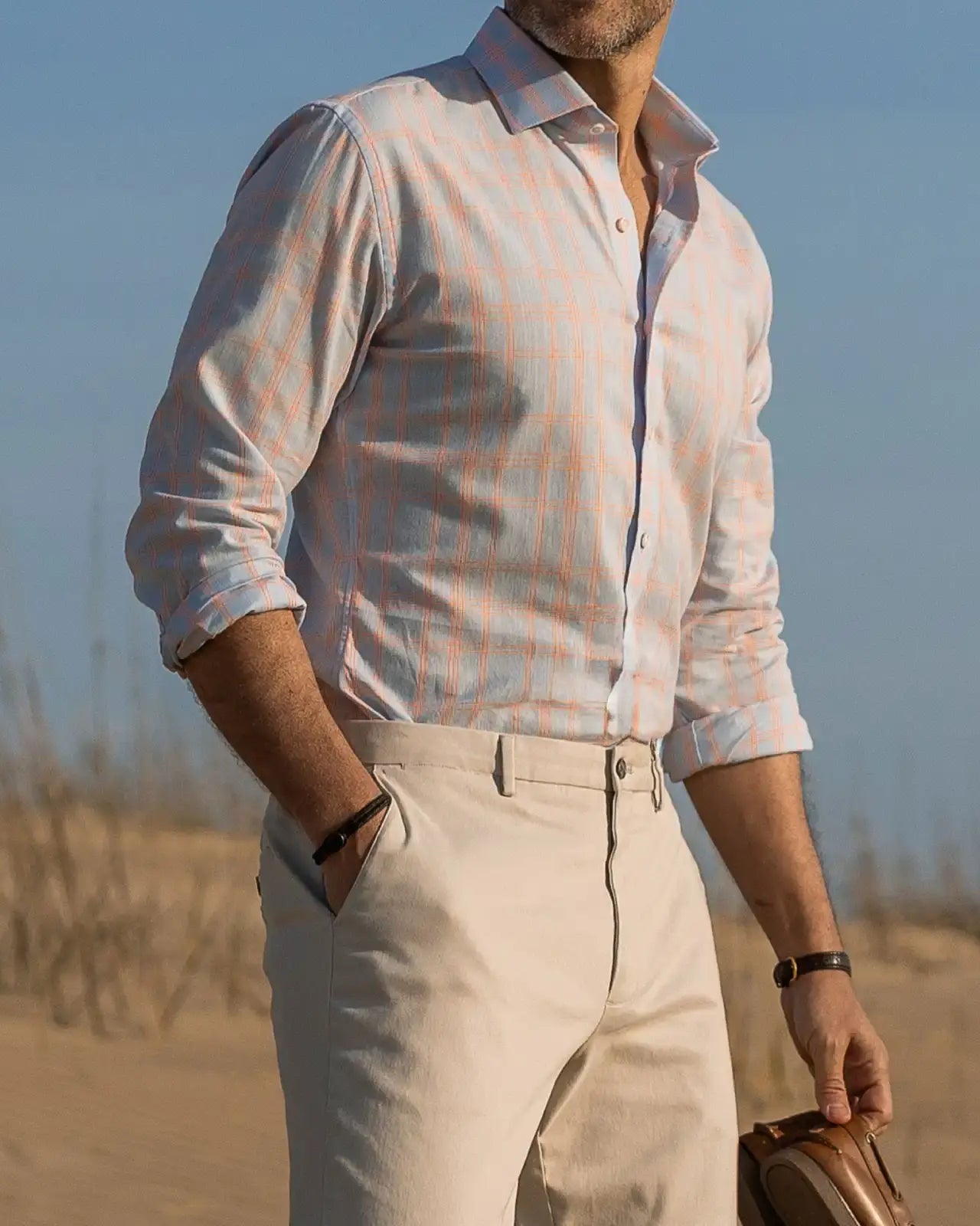 Man standing on a beach wearing a light blue and bright orange checkered shirt and tan chinos