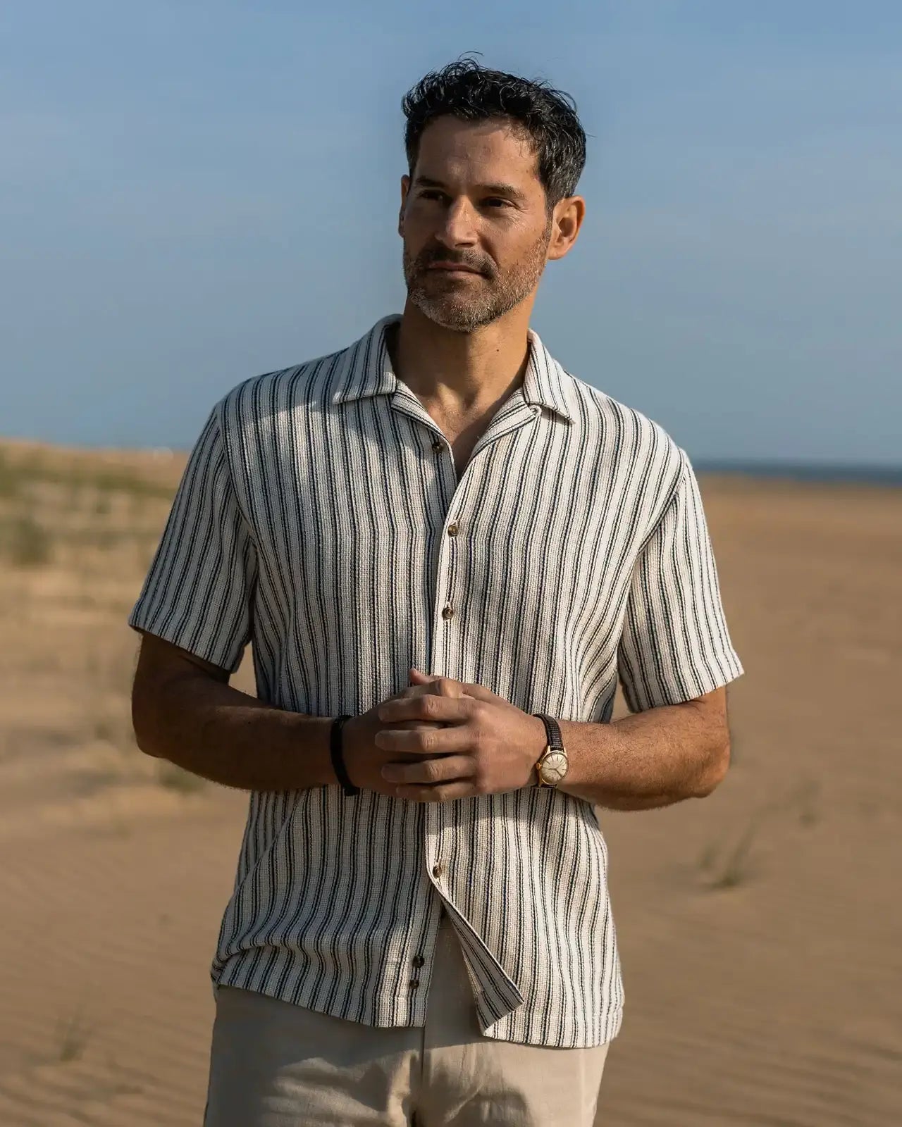 Man standing in on the beach wearing a striped short sleeve camp collar shirt, shorts and watch.