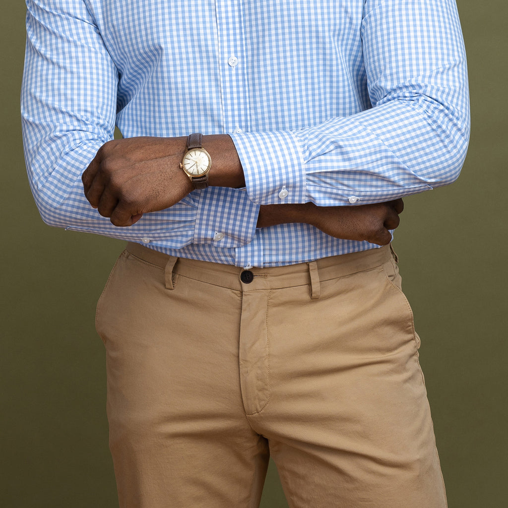 Close up on man wearing tan khaki pants and blue & white dress shirt in front of an olive green backdrop.