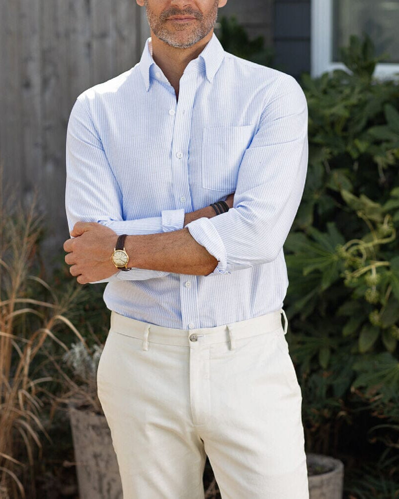 Man wearing a light blue striped shirt and beige pants standing outdoors with plants in the background
