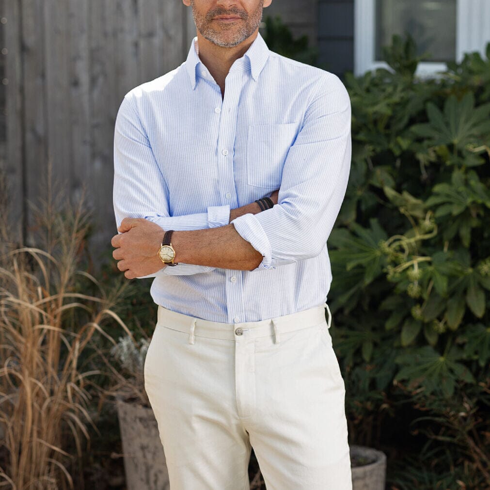 Man wearing a light blue striped shirt and beige pants standing outdoors with plants in the background