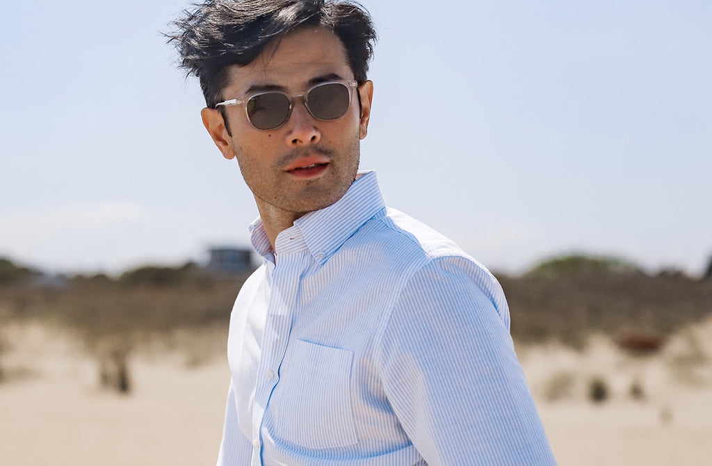 Photo of a model walking on a beach wearing a striped blue and white oxford button down shirt and sunglasses.