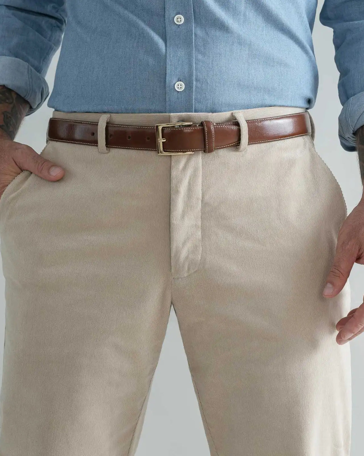 A close-up of a person wearing tan corduroy chinos with a brown leather belt and a light blue button-down shirt, cropped from the chest down against a light gray background.