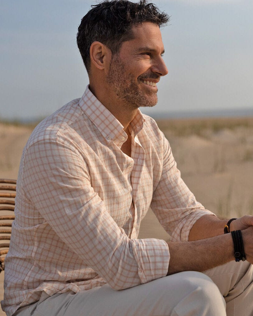 Model sitting down in a beach setting wearing a peach colored checked cotton linen shirt by Ledbury