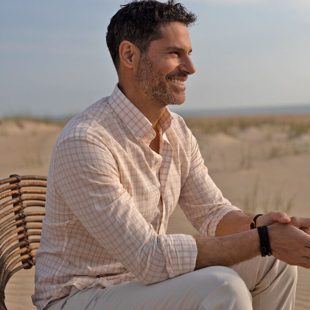 Model sitting down in a beach setting wearing a peach colored checked cotton linen shirt by Ledbury
