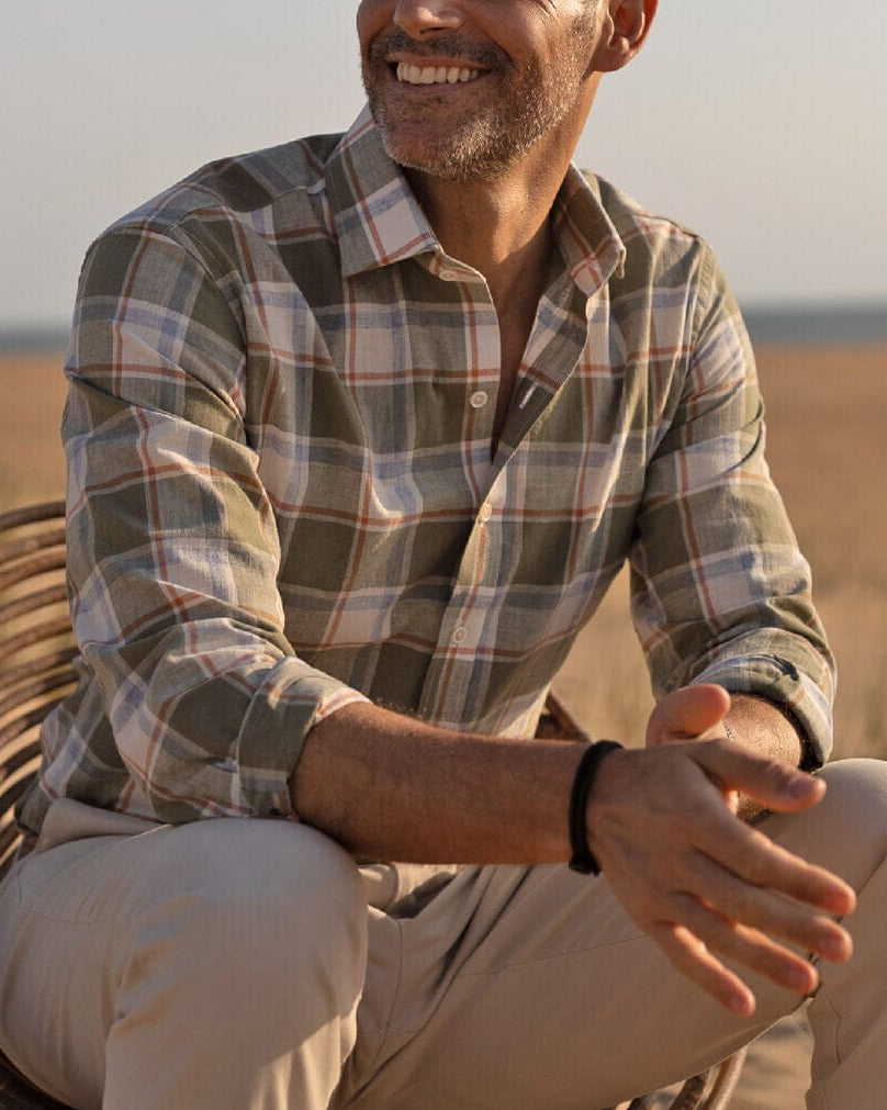 Man sitting outdoors wearing a plaid shirt and beige pants, with a blurred natural background.