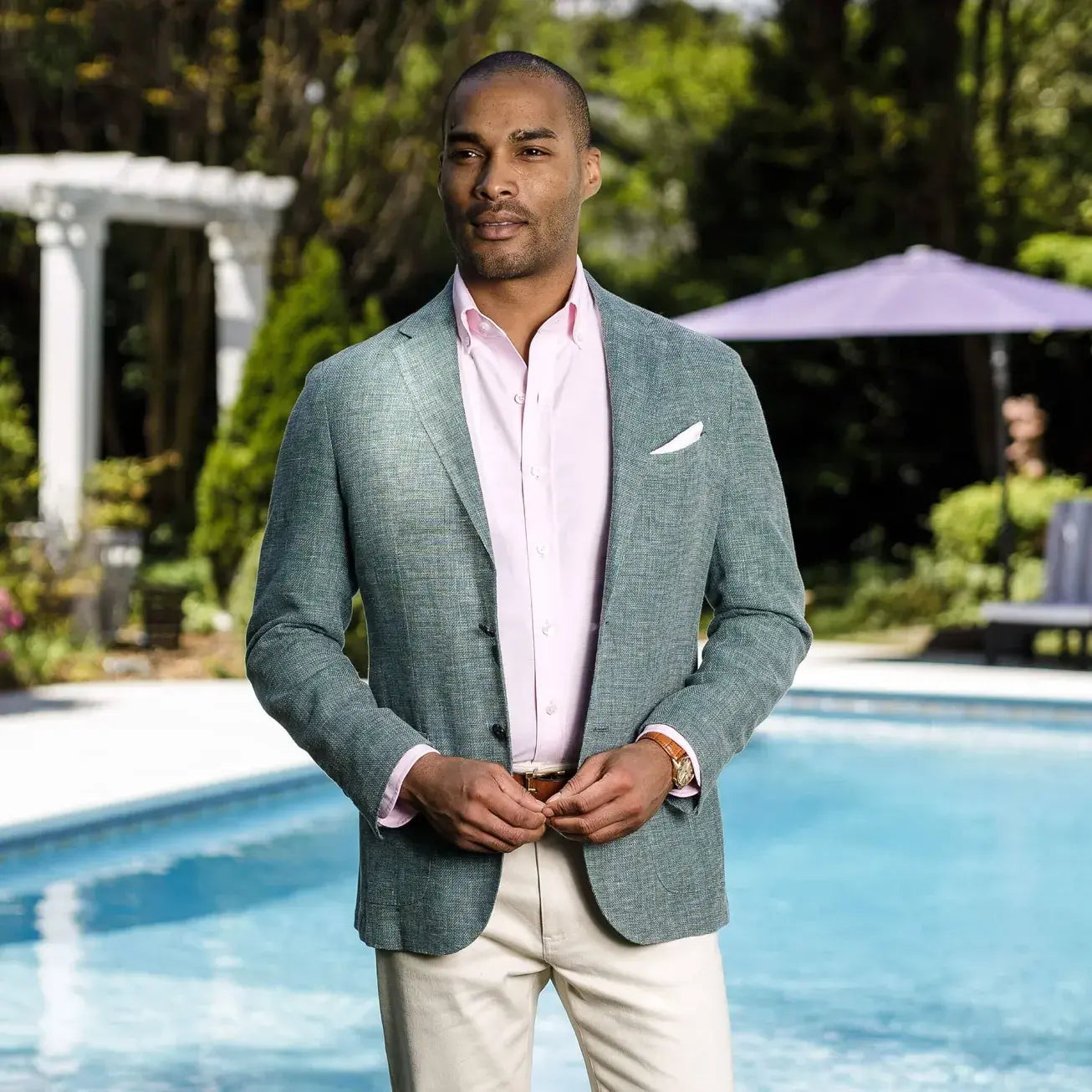 Male model wearing a light pink Ledbury button down shirt with an emerald green blazer. He is standing outside in a lush environment, in front of a swimming pool.