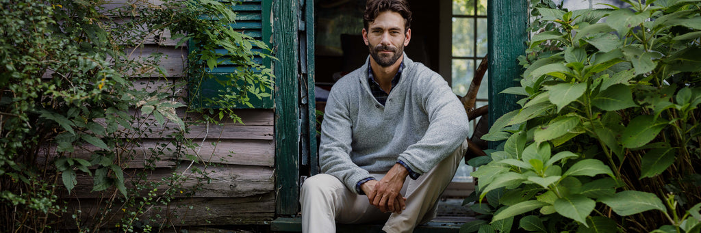 Image of a man wearing a light grey crewneck sweater. He is sitting on the steps of a rustic house surrounded by greenery. 