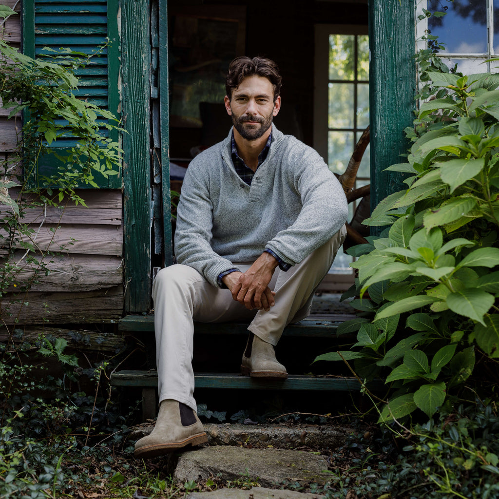 Image of a man wearing a light grey crewneck sweater. He is sitting on the steps of a rustic house surrounded by greenery. 