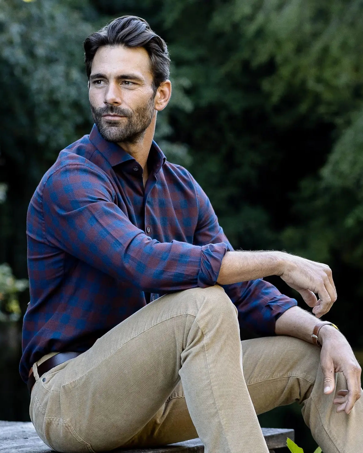 A man sitting outdoors by a body of water wearing a blue and red plaid button-down shirt and tan pants, with a lush green forest background.