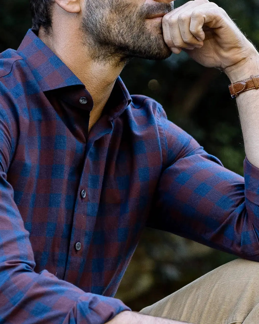 A close-up lifestyle shot of a man wearing a long-sleeve plaid button-down shirt in dark red and blue, paired with khaki pants and a brown leather watch.