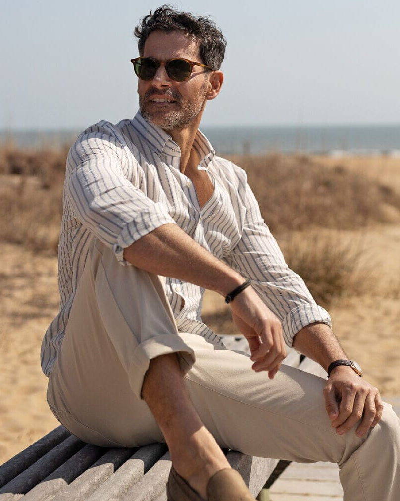 Man sitting on a wooden bench by the beach wearing sunglasses and a light blue striped cotton linen shirt