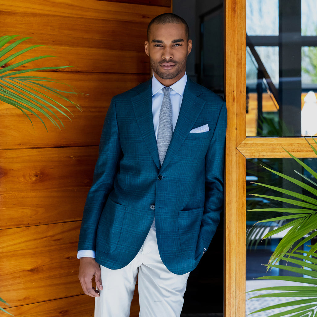 Male model wearing a blue dress shirt, tie, and teal green sport coat. He is standing in a wooden doorway with palm plants around. 
