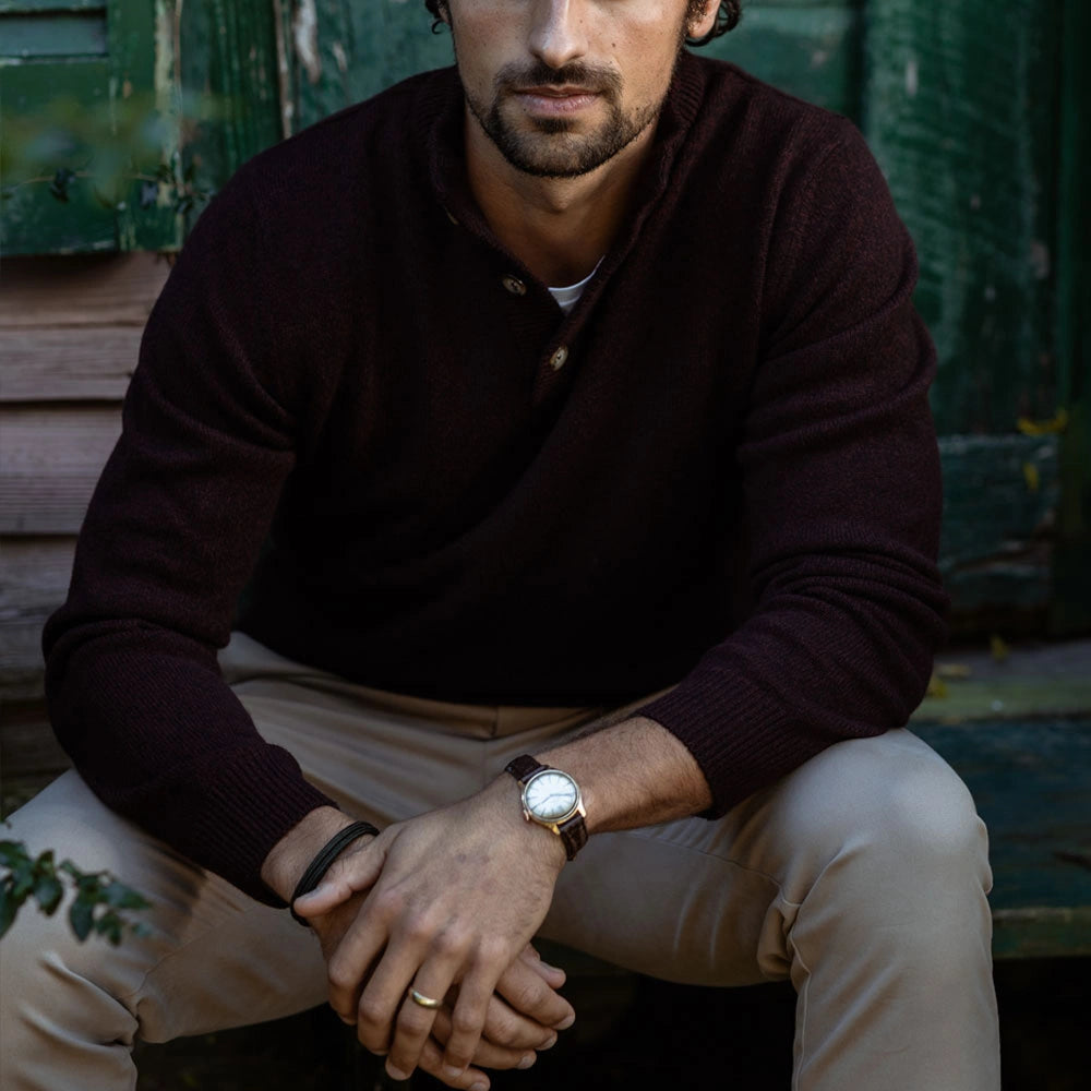 A man wearing a dark burgundy knit henley sweater and beige trousers, sitting in front of a rustic green wooden door. He is wearing a brown leather watch with a white dial on his wrist.