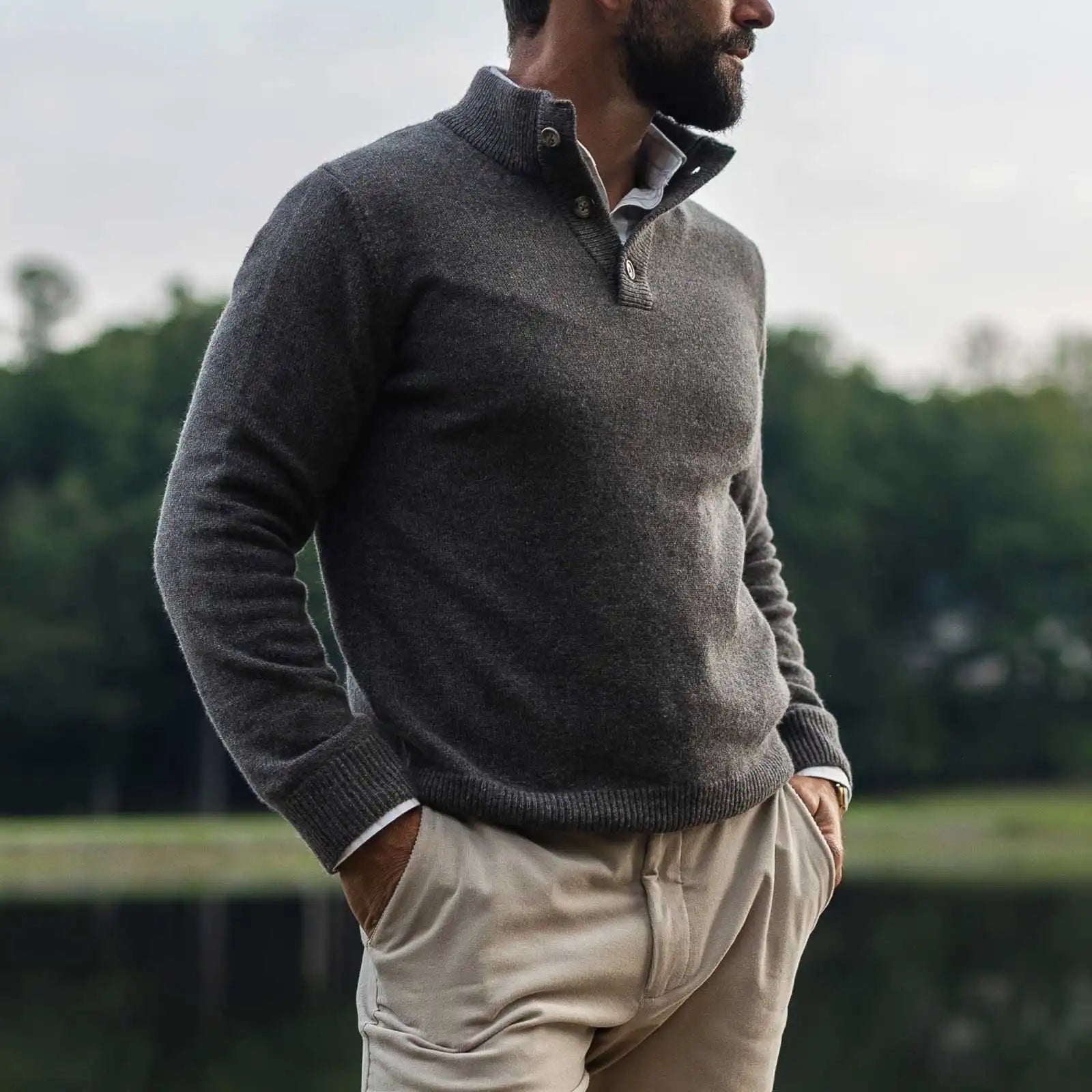 Man wearing a taupe light brown mock neck Italian merino sweater. He is standing outside in front of a lake with a blurred background. 