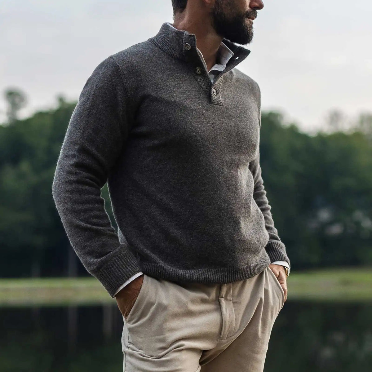 Man wearing a taupe light brown mock neck Italian merino sweater. He is standing outside in front of a lake with a blurred background. 