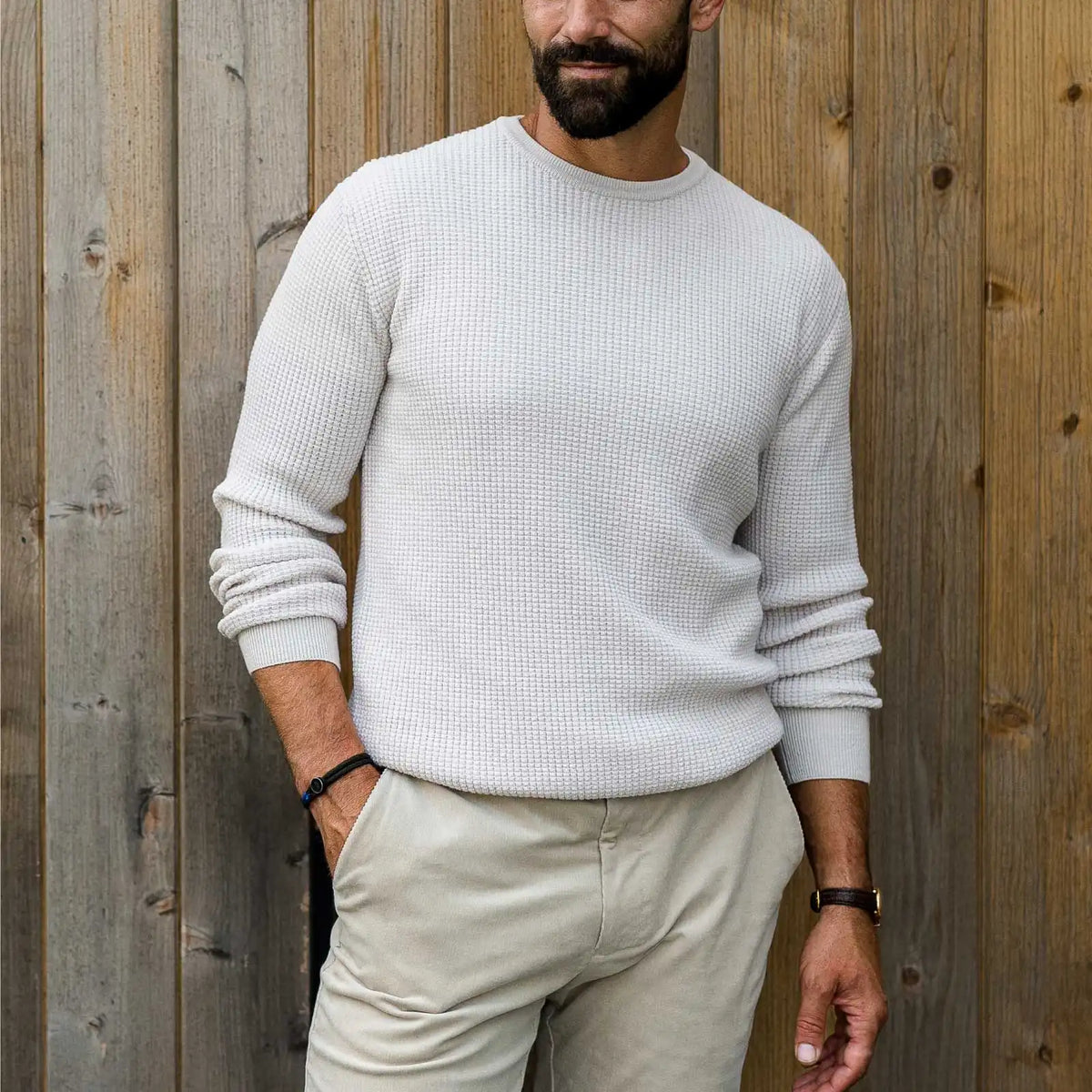 Man wearing a cream crew neck, waffle knit merino sweater by Ledbury. He is standing in front of a wood slat wall outdoors. 
