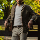 A man wearing a dark olive green waxed cotton jacket over a white ribbed knit sweater, paired with beige trousers and a brown belt, leaning against a wooden fence in an outdoor park setting.