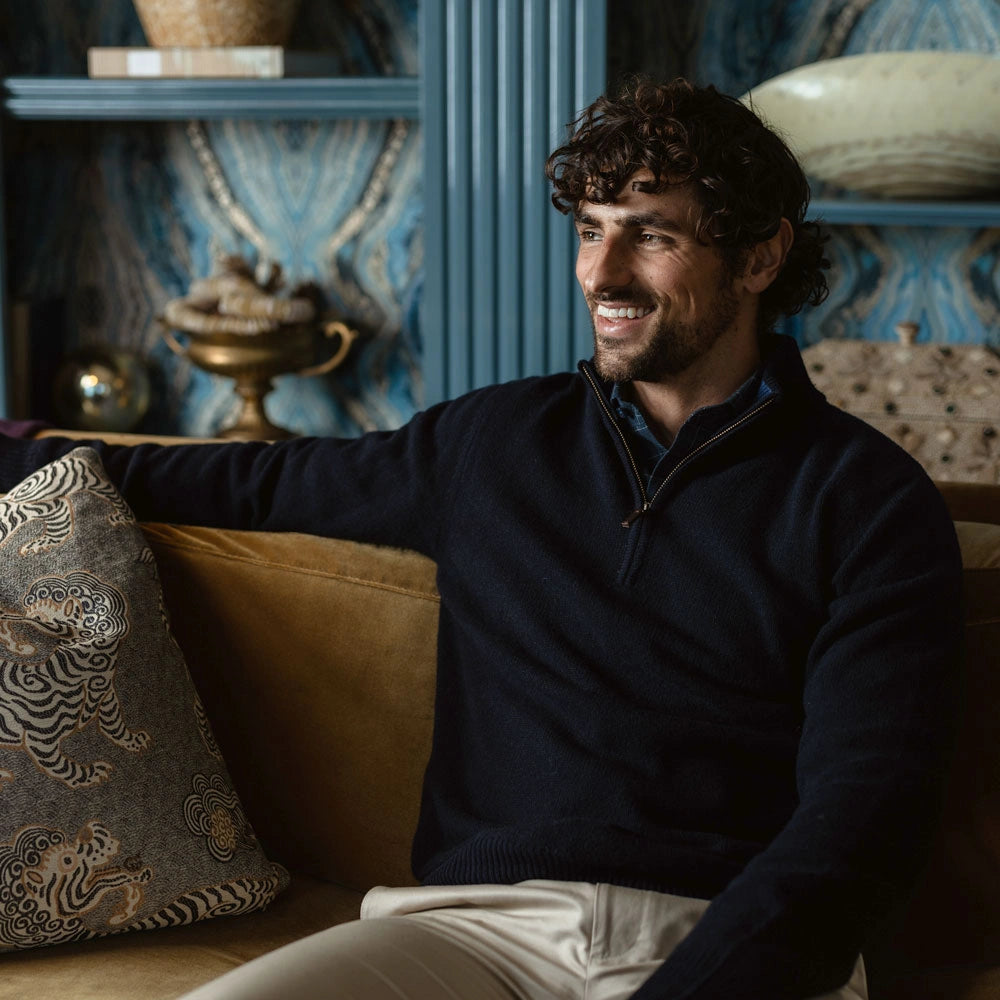 A smiling man with curly dark hair sits on a tan sofa wearing a dark navy quarter-zip sweater and light beige trousers, with a decorative pillow beside him in an ornate blue and gold room.