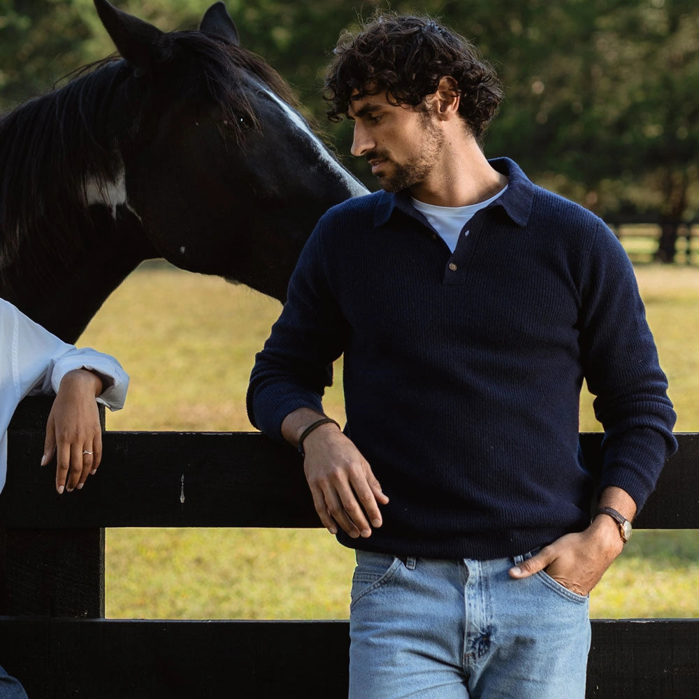 A man wearing a navy blue knit polo sweater and light blue jeans stands outdoors next to a black fence, with a horse visible behind him. The product being shown is the navy sweater.